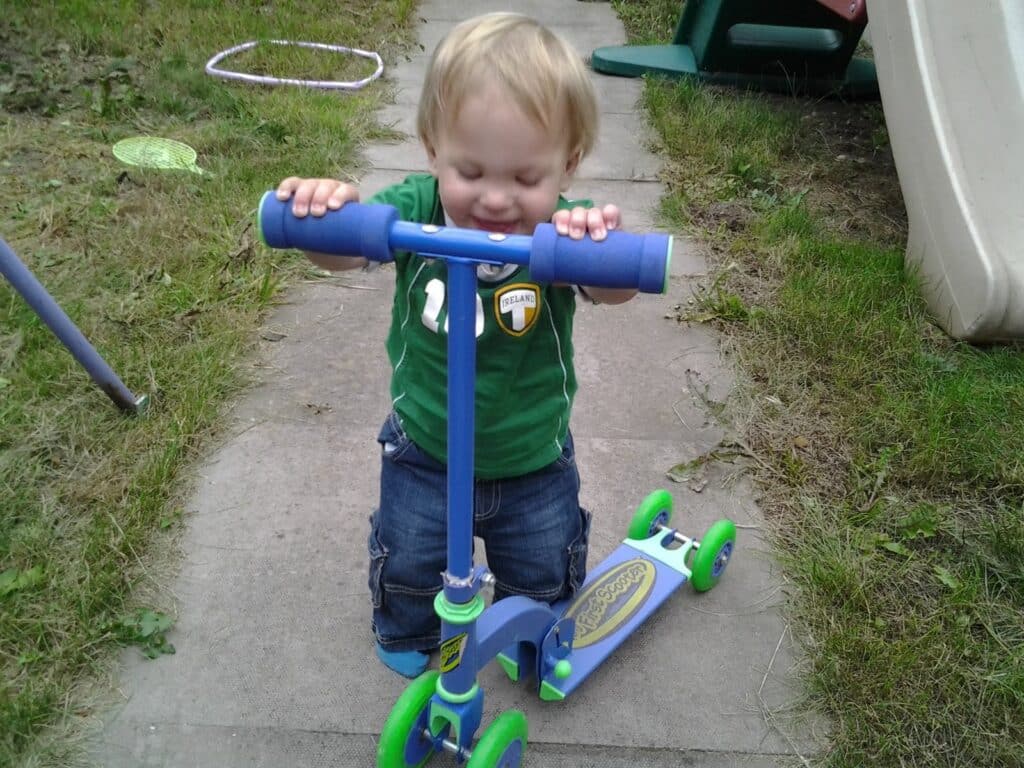 A toddler with blond hair, wearing a green shirt and blue shorts, kneels on a path outdoors, holding the handlebars of a blue and green scooter. Grass and playground equipment are visible in the background. The child is smiling with eyes closed.