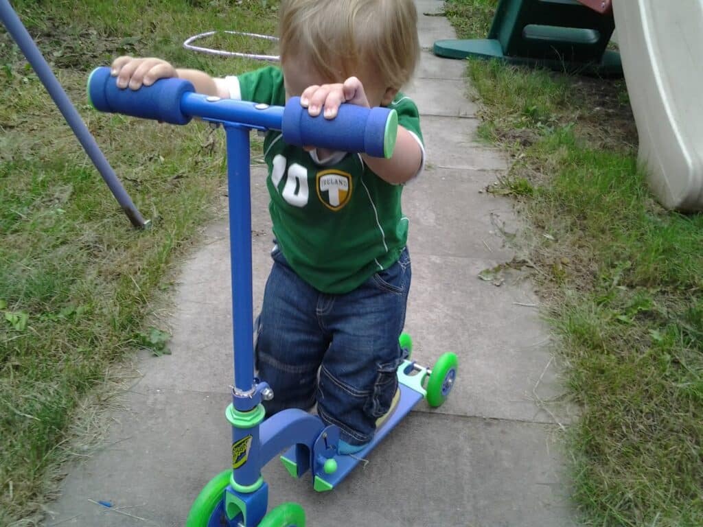A young child with blond hair wearing a green Ireland t-shirt and blue jeans is kneeling on a blue and green scooter on a paved garden path. The child is holding both handlebars and looking down, with grass and playground equipment in the background.