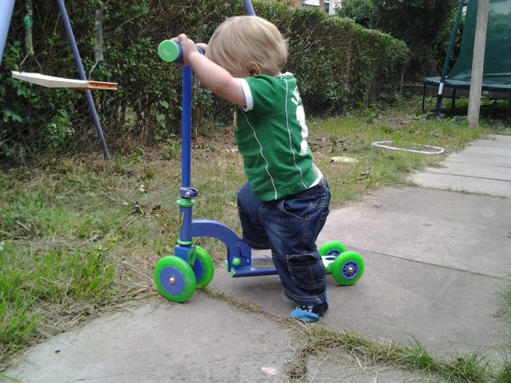 A toddler with blond hair, wearing a green shirt and blue jeans, stands on a stone path in a garden, holding the handlebars of a blue and green three-wheeled scooter. The child faces away, and there is grass, a swing set, and a trampoline nearby.