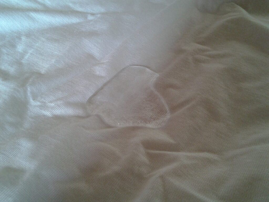 A small, transparent wet spot is visible on a smooth white fabric surface. The surrounding fabric appears clean and uncreased, drawing attention to the irregularly shaped damp patch in the centre.