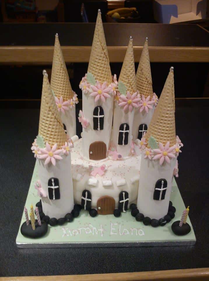 A castle-shaped cake decorated with ice cream cones as towers, topped with pink flowers and white pearls. The cake has white icing, small doors and windows, and “Xiorant Elana” written in light blue icing at the base. Three lit candles are in front.