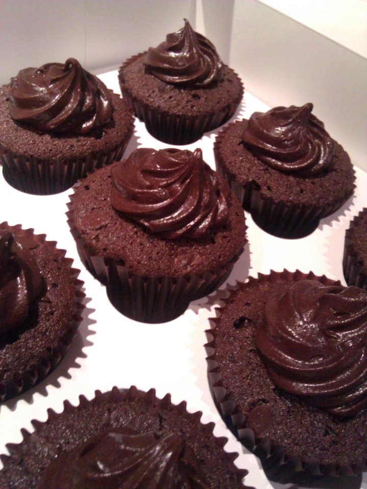 A box of eight chocolate cupcakes with dark chocolate icing swirled on top of each, displayed in black paper cases. The cupcakes are arranged closely together on a white surface inside a box, emphasising their rich, glossy appearance.