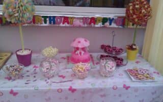 A pastel-themed birthday dessert table features a pink cake with a horse and mushrooms centrepiece, jars of sweets, bowls of confectionery, and tree-shaped sweet displays. A colourful “Happy Birthday” banner hangs above the table, which is covered with a butterfly-patterned cloth.