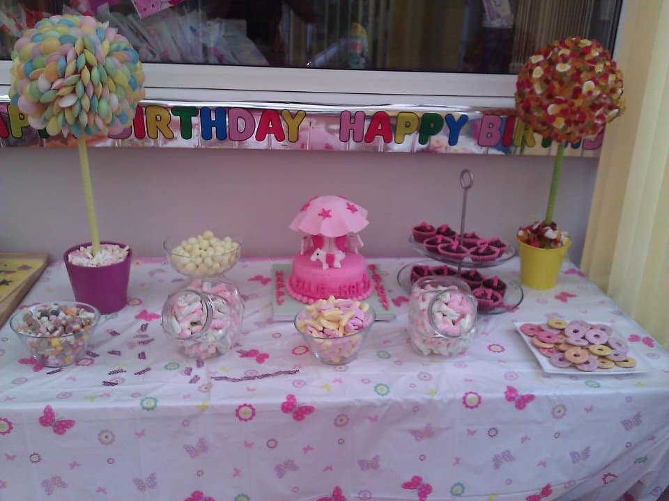 A pastel-themed birthday dessert table features a pink cake with a horse and mushrooms centrepiece, jars of sweets, bowls of confectionery, and tree-shaped sweet displays. A colourful “Happy Birthday” banner hangs above the table, which is covered with a butterfly-patterned cloth.