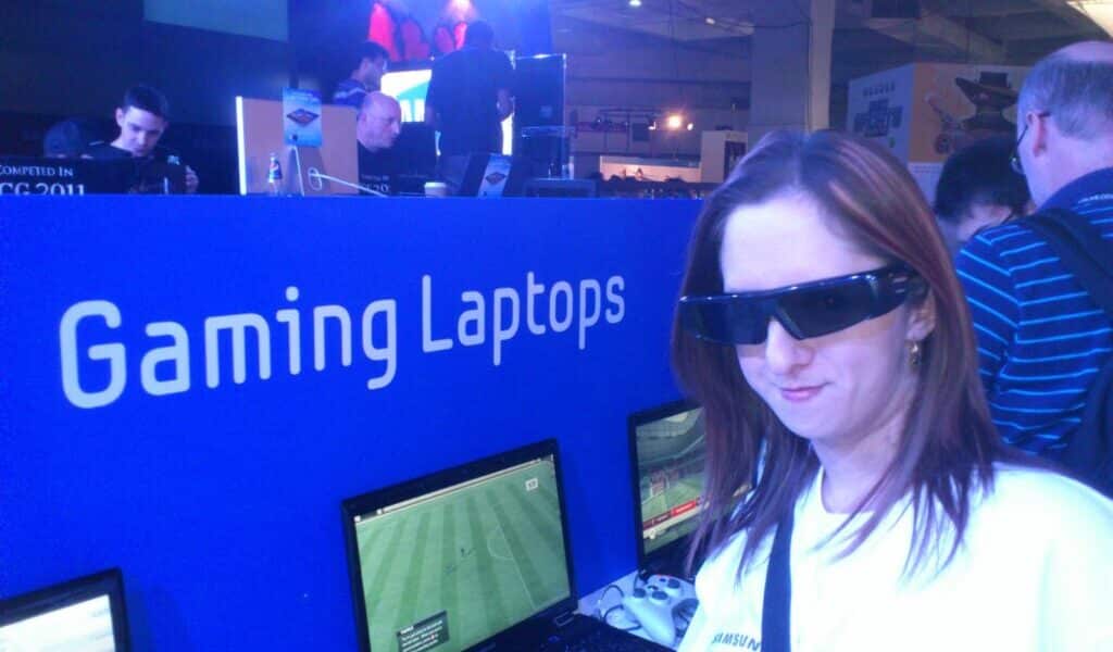 A woman wearing 3D glasses stands in front of a row of gaming laptops displaying a football video game. Behind her, a blue stand labelled "Gaming Laptops" is visible, with people using laptops and stage lights shining in the background.