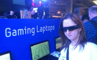 A woman wearing 3D glasses stands in front of a row of gaming laptops displaying a football video game. Behind her, a blue stand labelled "Gaming Laptops" is visible, with people using laptops and stage lights shining in the background.