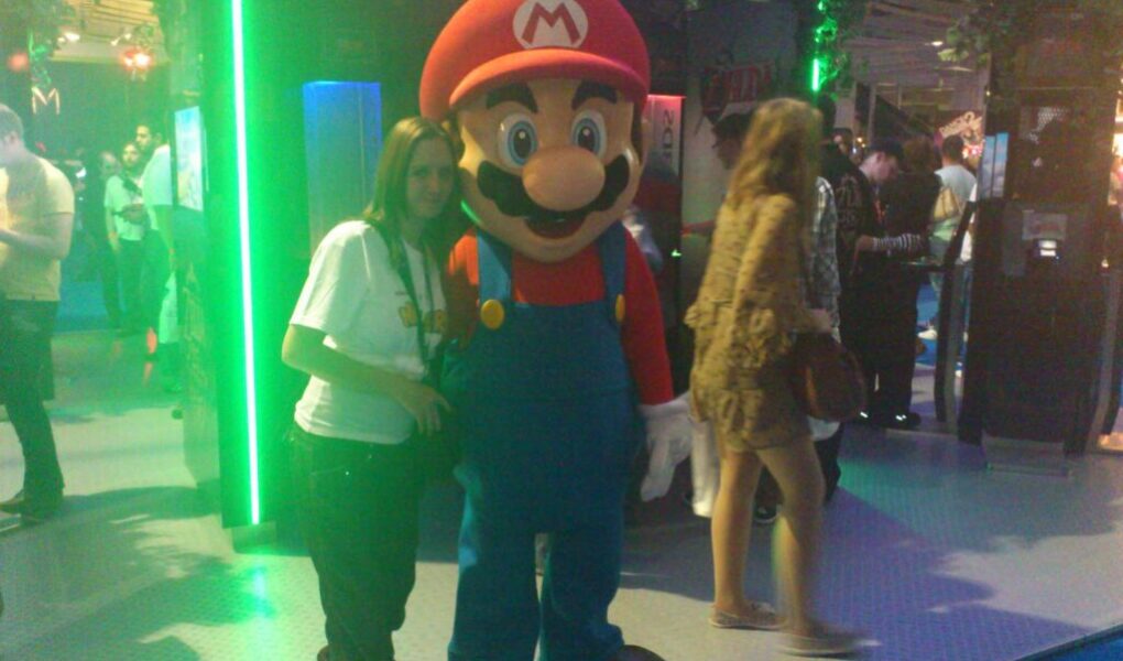 A person poses next to a life-sized Mario mascot in a convention setting, surrounded by other attendees and green-lit displays. The area is lively with people walking and interacting, and there is lush plant decor above the stands.