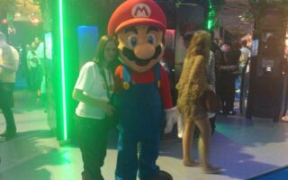 A person poses next to a life-sized Mario mascot in a convention setting, surrounded by other attendees and green-lit displays. The area is lively with people walking and interacting, and there is lush plant decor above the stands.