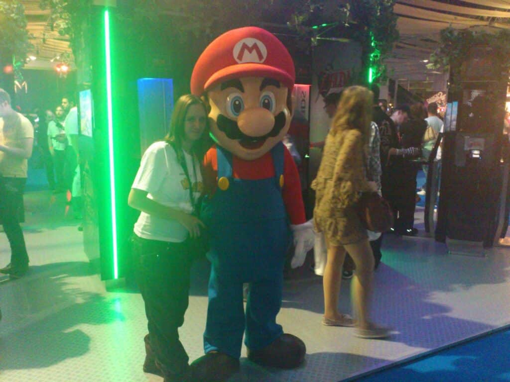 A person poses next to someone in a large Mario costume at an indoor event with neon lighting. Other people walk by or stand in the background near stands and gaming setups. The setting is lively and colourful, with a playful, festive atmosphere.