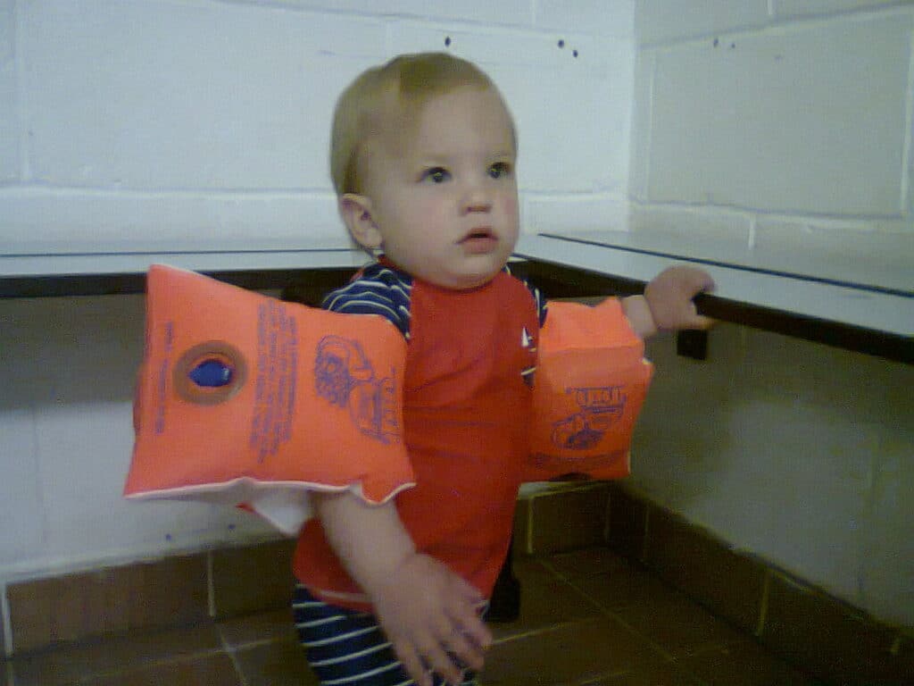 A toddler with short, light brown hair and a serious expression stands indoors. The child is wearing red and blue clothing and has bright orange inflatable swimming armbands on both arms. The background features white tiled walls and a tiled floor.