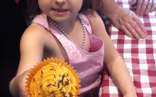 A young girl in a pink dress and tiara holds out a yellow-frosted cupcake with chocolate sprinkles. She stands at a red and white chequered table, with an adult’s hand resting on the table beside her. The girl looks proud and is smiling slightly.