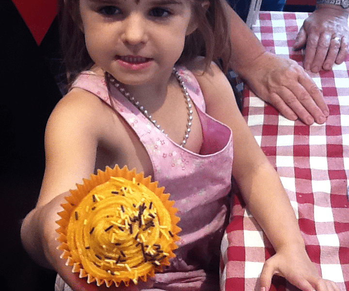 A young girl in a pink dress and tiara holds out a yellow-frosted cupcake with chocolate sprinkles. She stands at a red and white chequered table, with an adult’s hand resting on the table beside her. The girl looks proud and is smiling slightly.