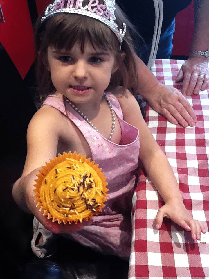 A young girl in a pink dress and tiara holds out a yellow-frosted cupcake with chocolate sprinkles. She stands at a red and white chequered table, with an adult’s hand resting on the table beside her. The girl looks proud and is smiling slightly.