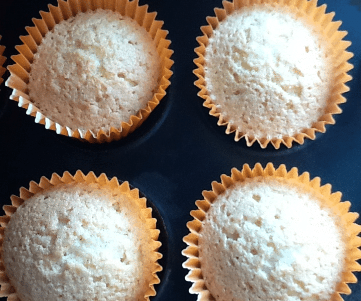 A close-up of six plain cupcakes in golden paper cases, arranged in a black muffin tray. The cupcakes have a light, golden-brown colour and a slightly domed, textured top. The background at the bottom is a turquoise surface.