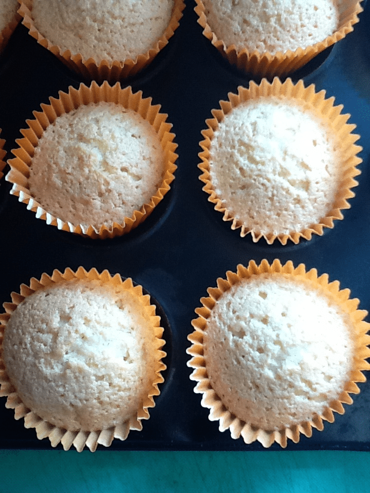 A close-up of six plain cupcakes in golden paper cases, arranged in a black muffin tray. The cupcakes have a light, golden-brown colour and a slightly domed, textured top. The background at the bottom is a turquoise surface.