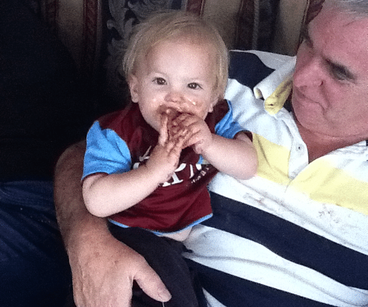 A smiling toddler with blonde hair and a maroon shirt sits on an older man's lap. The toddler’s hands and mouth are messy with chocolate. The man, wearing a striped polo shirt, looks at the child affectionately whilst sitting on a patterned sofa.