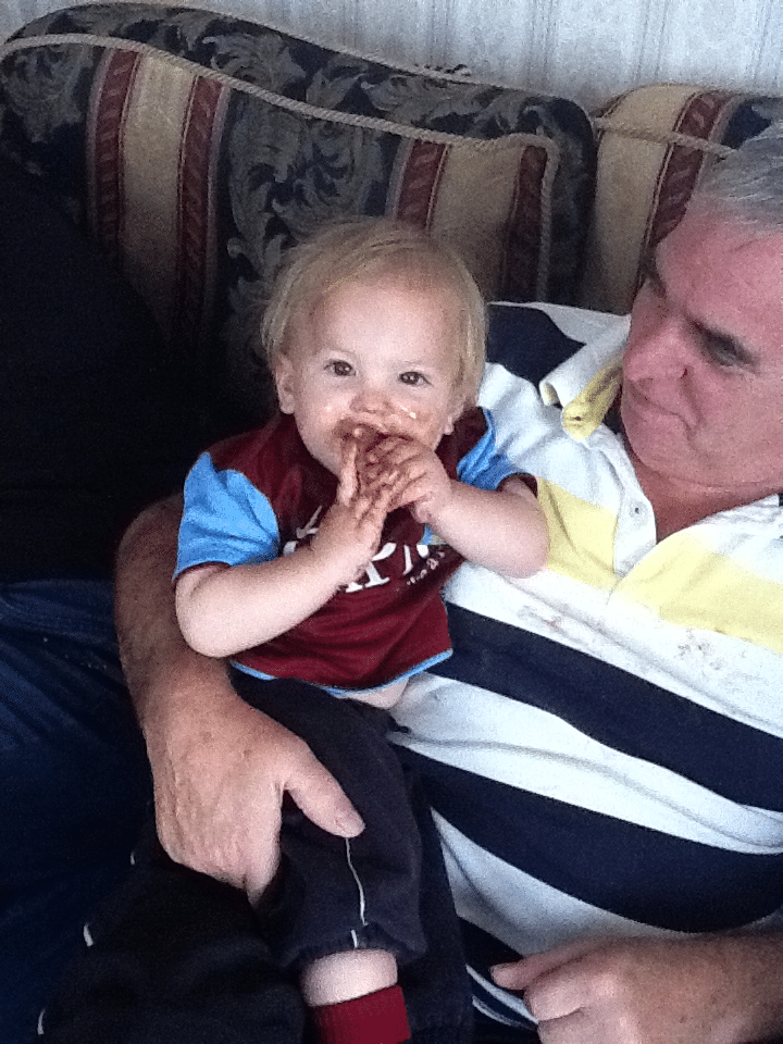 A smiling toddler with blonde hair and a maroon shirt sits on an older man's lap. The toddler’s hands and mouth are messy with chocolate. The man, wearing a striped polo shirt, looks at the child affectionately whilst sitting on a patterned sofa.