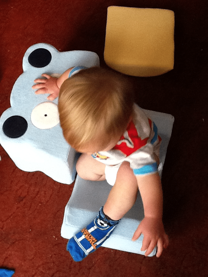 A toddler with blonde hair, wearing a white shirt with blue and red stripes and blue cartoon socks, sits sideways on a light blue armchair shaped like a bear, viewed from above. A small yellow cushion lies on the reddish-brown carpet nearby.
