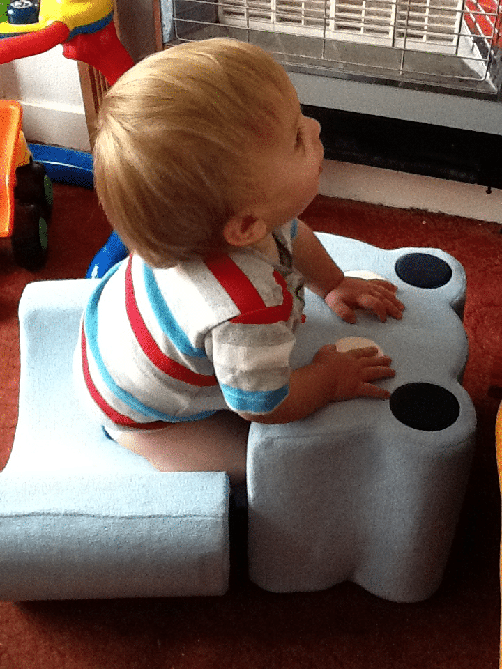 A young child with fair hair, wearing a striped shirt, sits sideways on a blue foam chair. The child is looking up and resting their hands on the chair’s arms. Toys and a metal heater are visible in the background on a red carpeted floor.