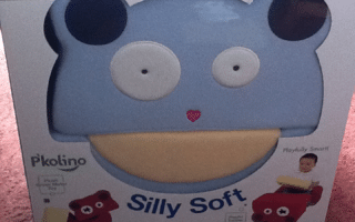 A box with a blue, bear-shaped foam seat called "Silly Soft" by P'kolino. The bear has round white eyes, dark blue ears, and a beige mouth area. The packaging shows a child sitting on the chair and includes the words "Playfully Smart.