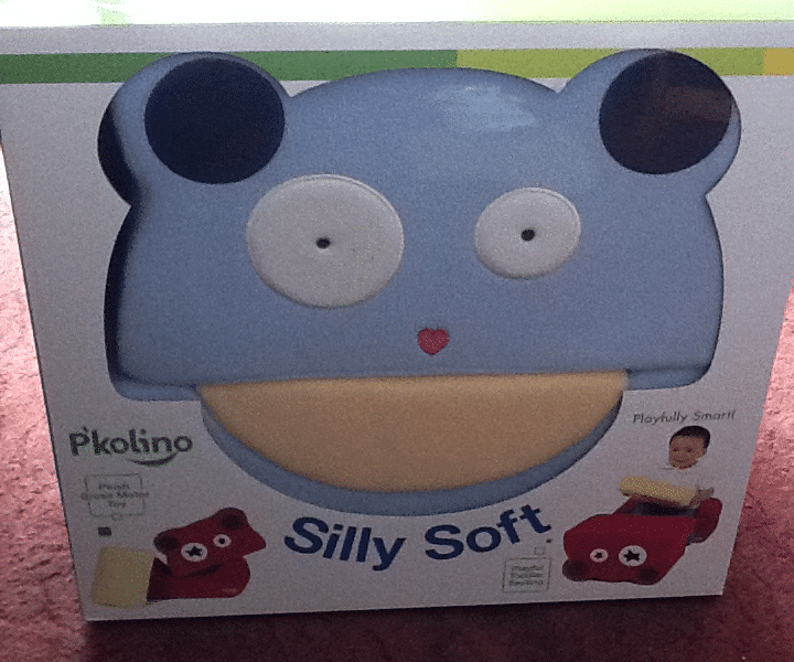 A box with a blue, bear-shaped foam seat called "Silly Soft" by P'kolino. The bear has round white eyes, dark blue ears, and a beige mouth area. The packaging shows a child sitting on the chair and includes the words "Playfully Smart.
