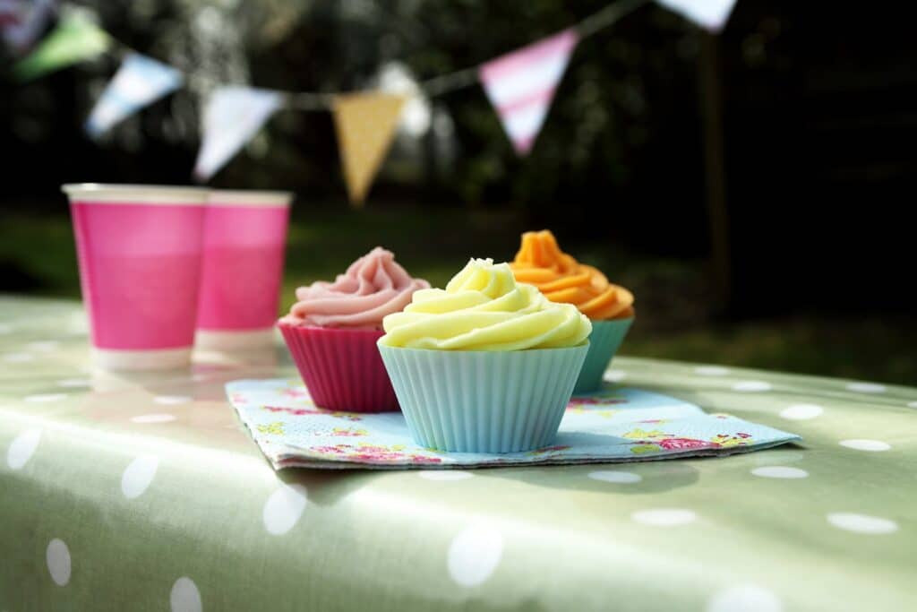 Three colourful cupcakes with pastel icing sit on a floral serviette atop a green spotty tablecloth. Two pink paper cups are nearby. In the blurred background, triangle bunting hangs, hinting at an outdoor celebration or picnic.