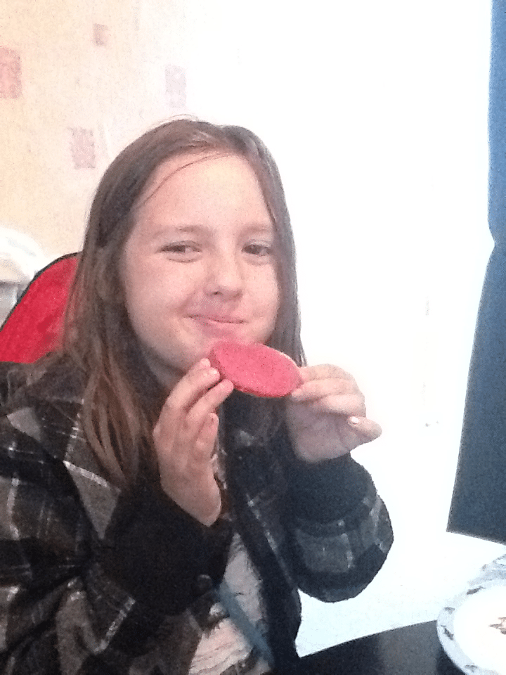A young child with long brown hair and a checked jacket is smiling whilst holding a slice of pink luncheon meat close to their mouth, sitting indoors at a table with a white plate partially visible in the foreground. The background is softly lit.
