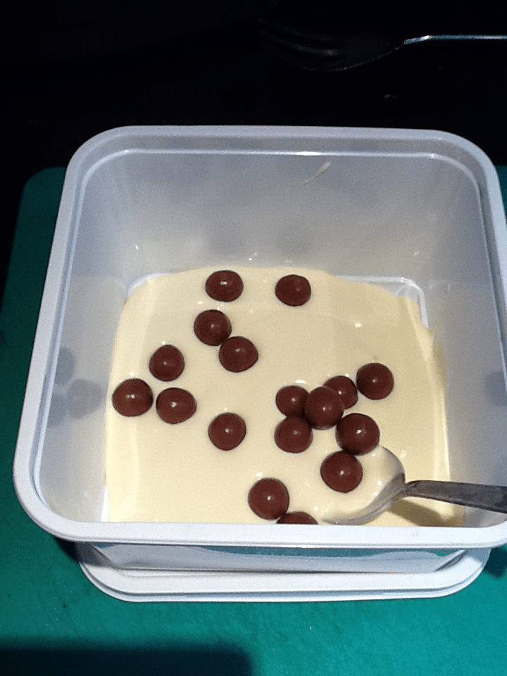 A white plastic container holding a layer of melted white chocolate with scattered round chocolate balls on top. A silver spoon rests in the corner of the container, and the container sits on a green surface. A fork is visible in the background.