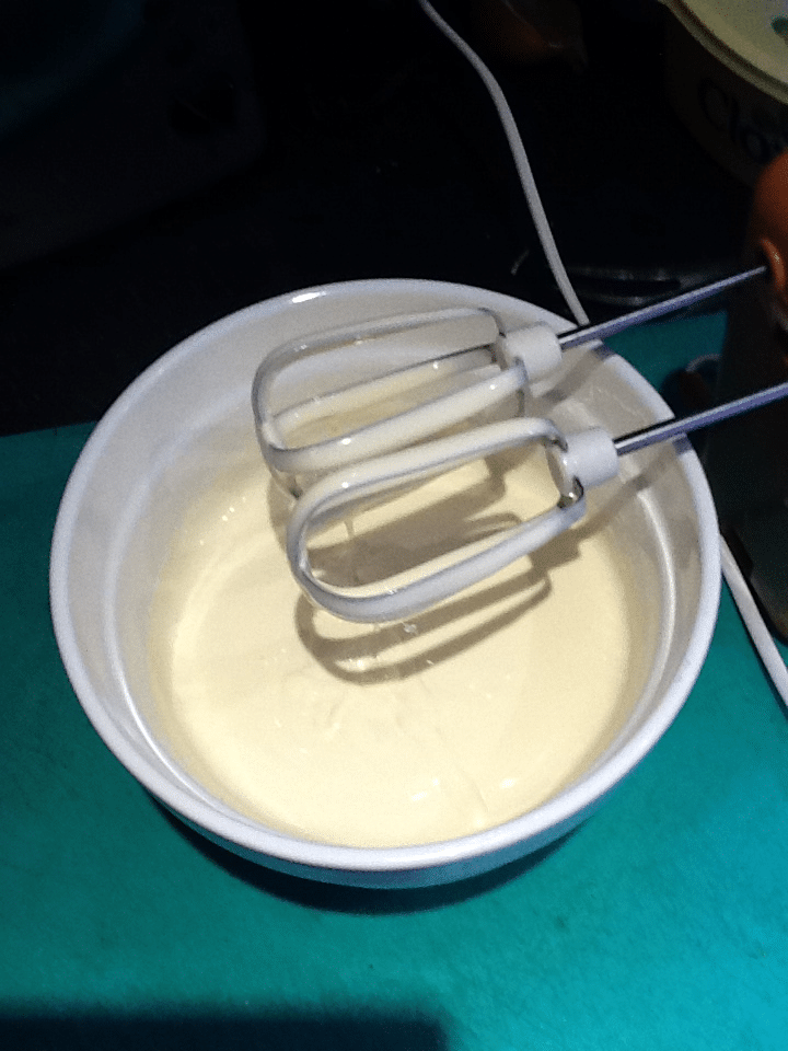 A white bowl filled with creamy, pale yellow mixture sits on a teal surface. Two metal beaters attached to a handheld electric whisk rest inside the bowl, suggesting the mixture has just been mixed. The background is dimly lit.