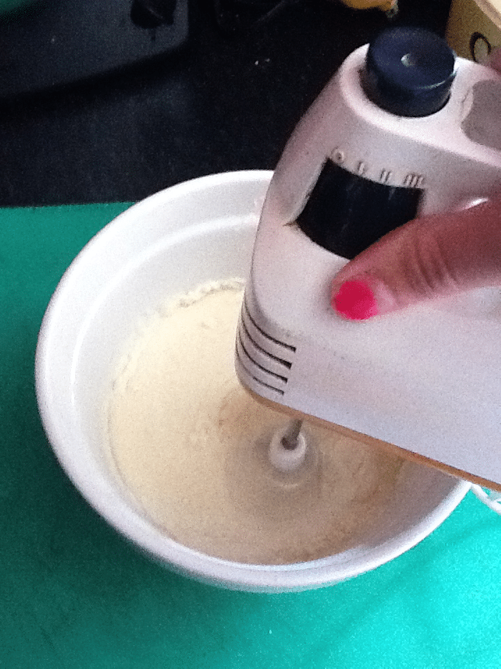 A hand with pink nail varnish holds a white electric mixer, blending a pale batter in a white bowl on a green surface. The mixer beaters are spinning, and the bowl is partially filled with the mixture.