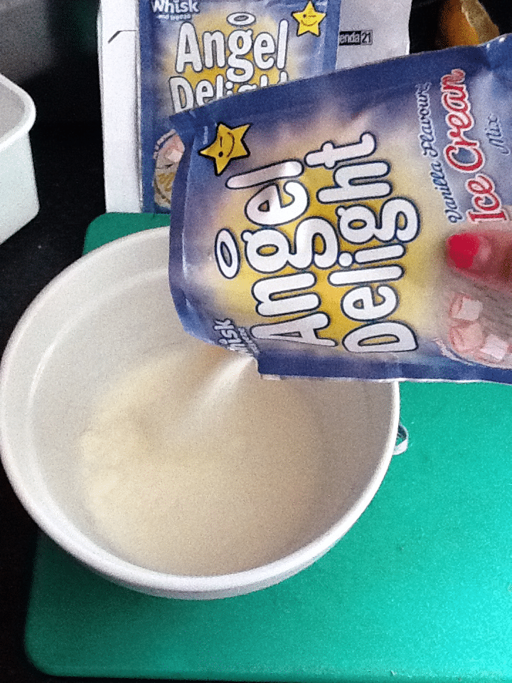 A hand pours powdered Angel Delight vanilla ice cream mix from a blue packet into a white bowl on a green surface. An opened packet and box of Angel Delight are visible in the background.