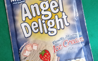 A blue sachet of Angel Delight Vanilla Flavour Ice Cream Mix lies on a green surface. The packaging features white and yellow text, a star cartoon, a strawberry image, ice cream swirls, and nutritional info at the bottom.