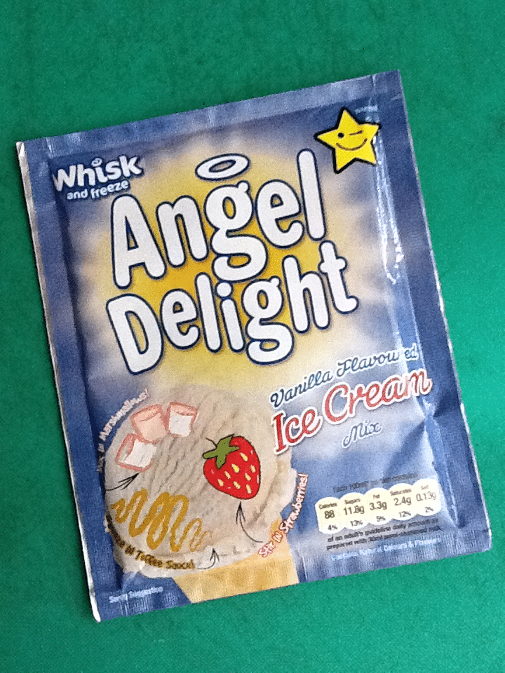 A blue sachet of Angel Delight Vanilla Flavour Ice Cream Mix lies on a green surface. The packaging features white and yellow text, a star cartoon, a strawberry image, ice cream swirls, and nutritional info at the bottom.