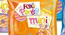 A close-up of a colourful package labelled "Fox's Party Mini Rings" shows playful, multicoloured text and fun doodles, including a crown and images of iced ring biscuits, on a bright orange background, suggesting a festive, cheerful snack.