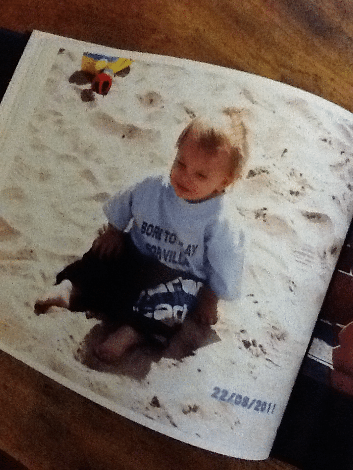 A toddler with blond hair sits on sand, wearing a light blue T-shirt and dark shorts. A small colourful toy is near their head. The photo appears in a printed album, with the date "22/08/2011" visible in blue text at the bottom right.