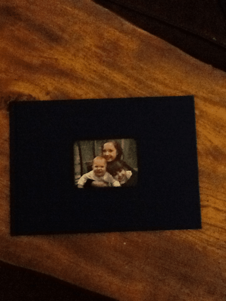 A small, slightly blurry photo of a smiling woman holding two young children is centred on a dark blue rectangular card placed on a wooden surface. The card contrasts with the warm brown wood underneath.