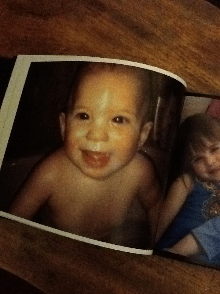 A printed photo album page shows a smiling baby with short dark hair, no shirt, and an open-mouthed grin. Next to the baby, a young girl in a blue dress appears partially in the frame. The album rests on a wooden surface.