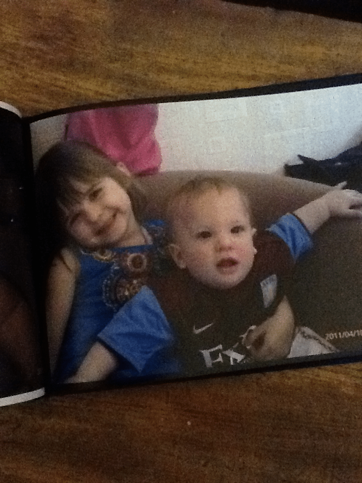 Two young children sit close together on a brown settee. The older girl, smiling, has straight brown hair and wears a blue top. The younger child is a baby in a maroon and blue football shirt, looking forward with one arm outstretched. A wooden surface is visible.