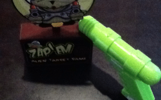 A small table-top game called "Zap! Alien Target Game" features a cartoon alien with three eyes on a red box. A bright green toy ray gun is pointed at the alien target, all set on a dark surface.