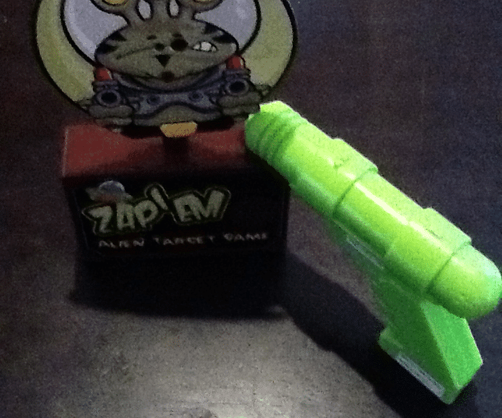 A small table-top game called "Zap! Alien Target Game" features a cartoon alien with three eyes on a red box. A bright green toy ray gun is pointed at the alien target, all set on a dark surface.
