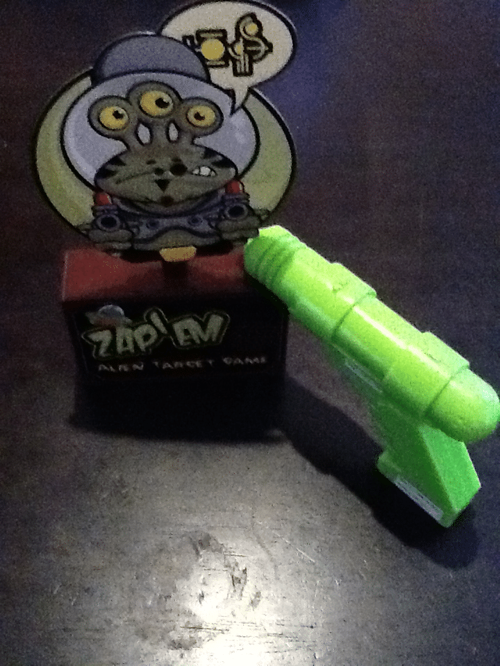 A small table-top game called "Zap! Alien Target Game" features a cartoon alien with three eyes on a red box. A bright green toy ray gun is pointed at the alien target, all set on a dark surface.