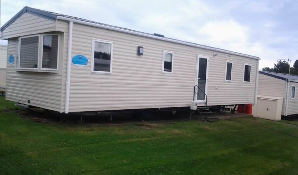 A beige mobile home sits on green grass under a cloudy sky. It has white-trimmed windows, a front door with steps and a handrail, and a blue sign near the front. A small shed and red bins are positioned near the rear of the home.