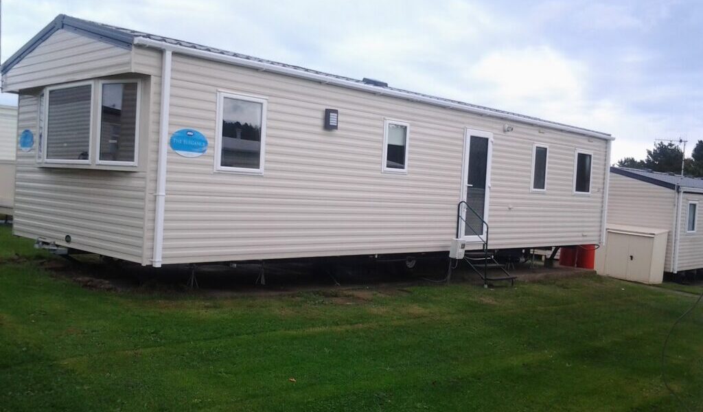 A beige mobile home sits on green grass under a cloudy sky. It has white-trimmed windows, a front door with steps and a handrail, and a blue sign near the front. A small shed and red bins are positioned near the rear of the home.