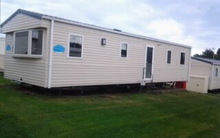 A beige mobile home sits on green grass under a cloudy sky. It has white-trimmed windows, a front door with steps and a handrail, and a blue sign near the front. A small shed and red bins are positioned near the rear of the home.