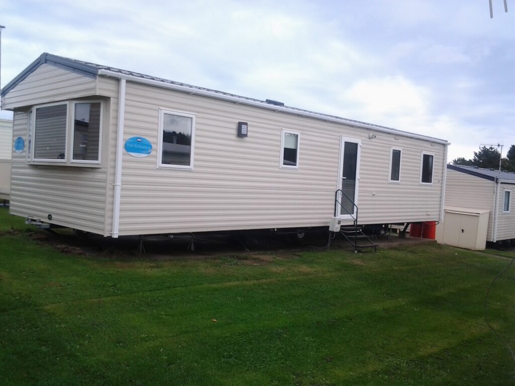 A beige mobile home with white trim is parked on a grassy area. It has several windows, a door with steps, and a blue oval sign near the front. A small shed and three red bins are visible in the background under a cloudy sky.