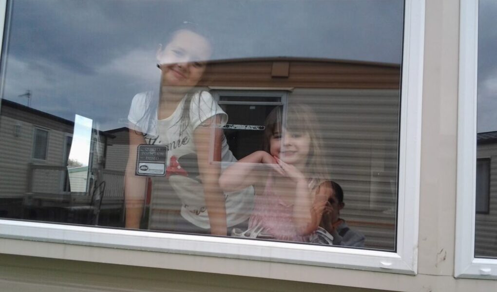 Two young girls smile and pose behind a large window, with a man partially visible taking the photo from inside. The window reflects a cloudy sky and nearby caravans. The girls appear cheerful, and the setting suggests a holiday park or mobile home area.