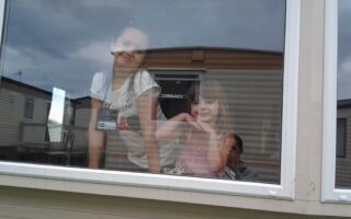 Two young girls smile and pose behind a large window, with a man partially visible taking the photo from inside. The window reflects a cloudy sky and nearby caravans. The girls appear cheerful, and the setting suggests a holiday park or mobile home area.