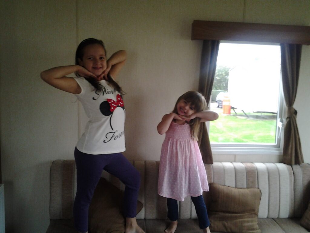 Two young girls stand barefoot on a beige-striped sofa, posing with hands under their chins and smiling. One girl wears a white Minnie Mouse top and purple trousers; the other wears a light pink dress. A window with brown curtains shows green grass outside.