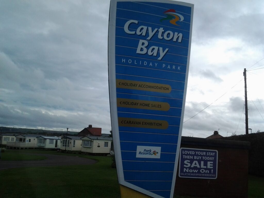 A large blue sign reads "Cayton Bay Holiday Park" with arrows to holiday accommodation, home sales, and exhibition. Below, a Park Resorts logo. Nearby, a sign advertises a sale. Static caravans and cloudy sky are in the background.