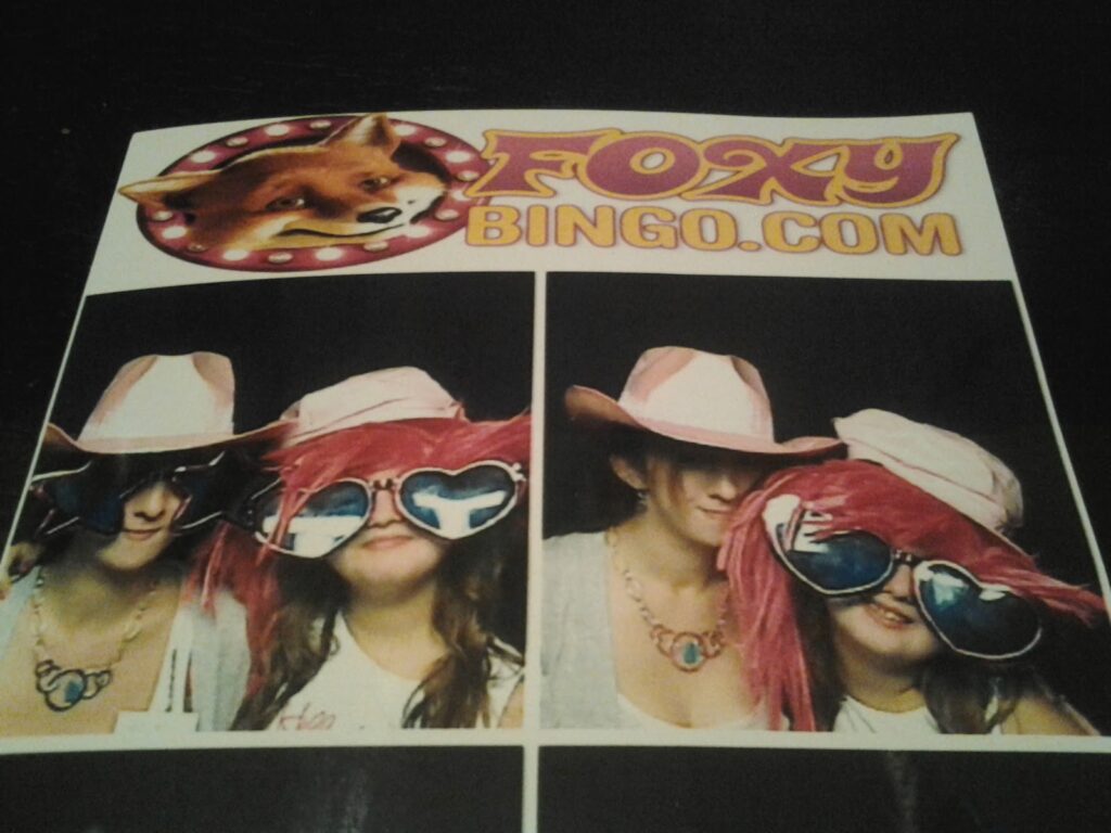 Two women wearing oversized sunglasses, hats, and costumes pose for a photo booth-style picture. The left panel shows them smiling, the right panel shows them closer together. Above is a "FoxyBingo.com" sign with a cartoon fox logo.